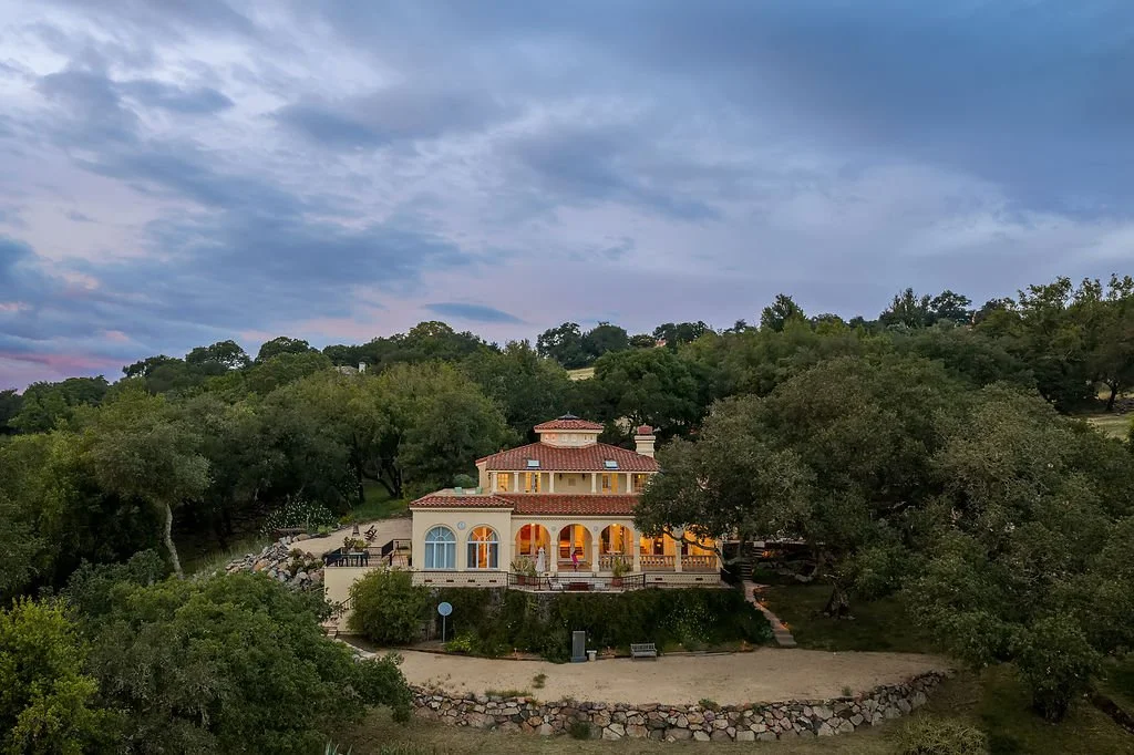 A house with a red tile roof and arched windows is surrounded by trees, with a sandy area and stone border in the foreground, under a partly cloudy sky.