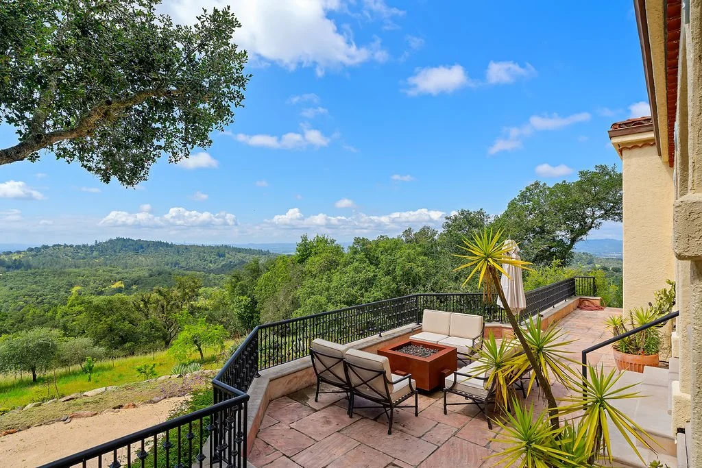 A spacious outdoor patio with beige seating around a fire pit, potted plants, and a view of green hills and blue sky with clouds.