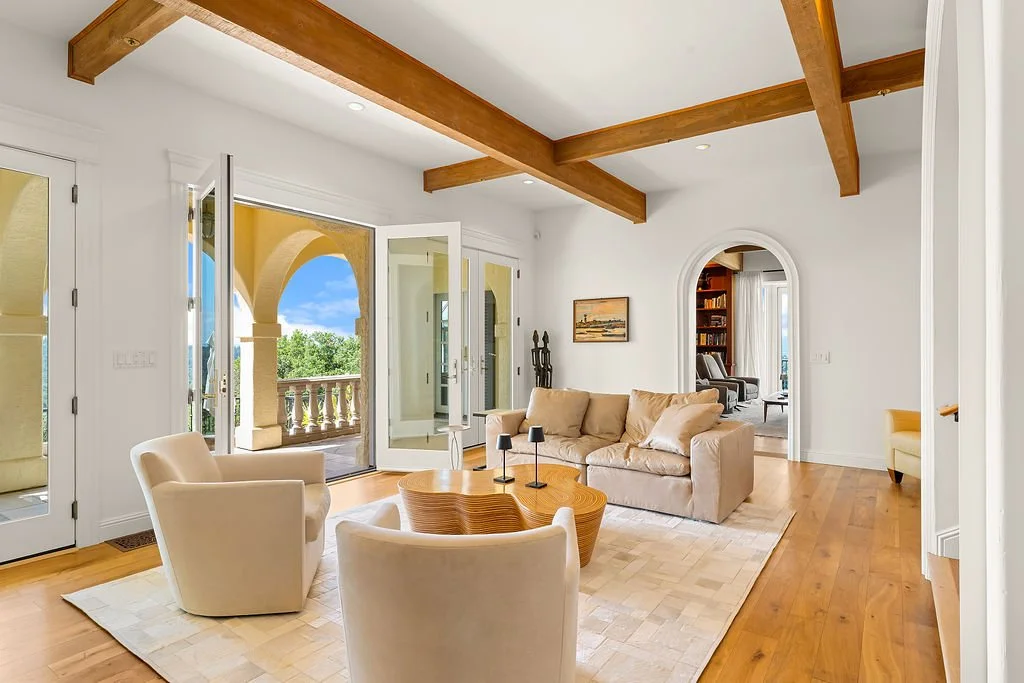 Living room with white walls, wooden ceiling beams, and hardwood floor, furnished with a beige sofa, three white armchairs, a wooden coffee table, and sliding glass doors opening to a balcony with arches and a view of green trees and blue sky.