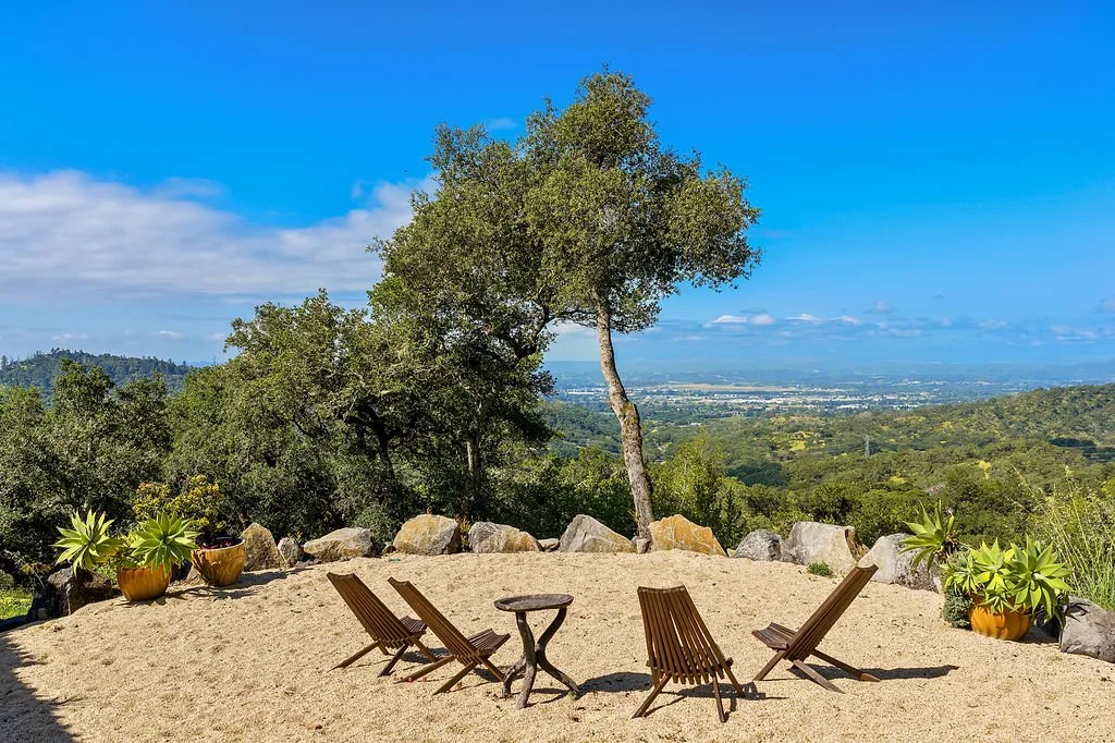 Outdoor patio with four wooden lounge chairs and a small table, surrounded by large rocks and potted plants, overlooking a scenic view of hills and a valley under a partly cloudy blue sky.