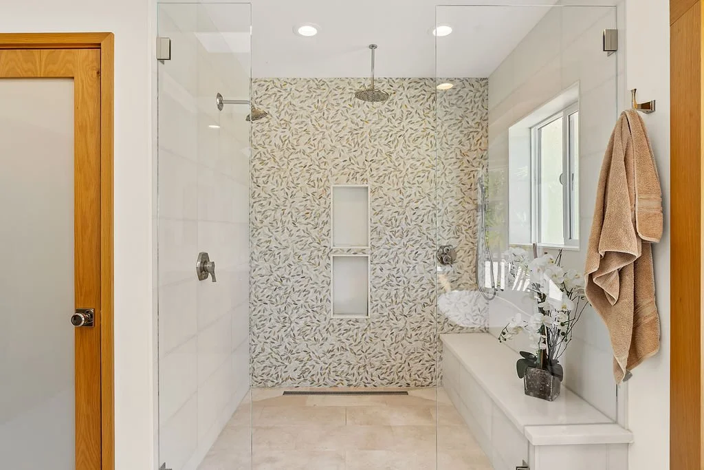 Modern walk-in shower with two rainfall shower heads, white built-in shelf, and a window, in a bathroom with light-colored floors and walls, beige towels, and a potted plant.