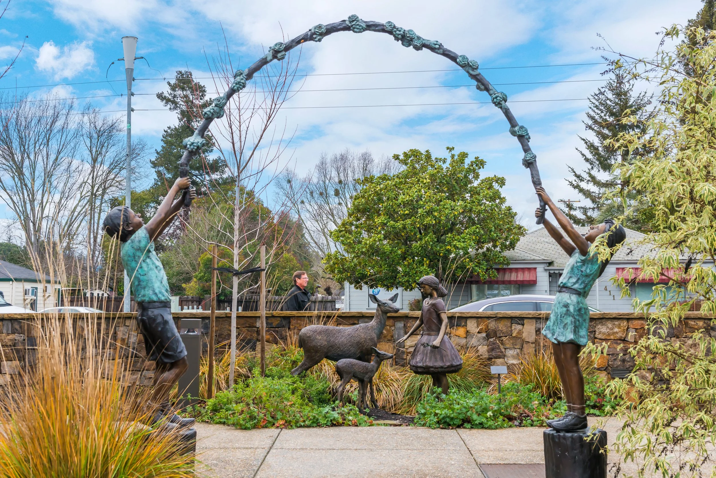 Outdoor sculpture of children holding a floral arch, with a deer sculpture and trees in the background.
