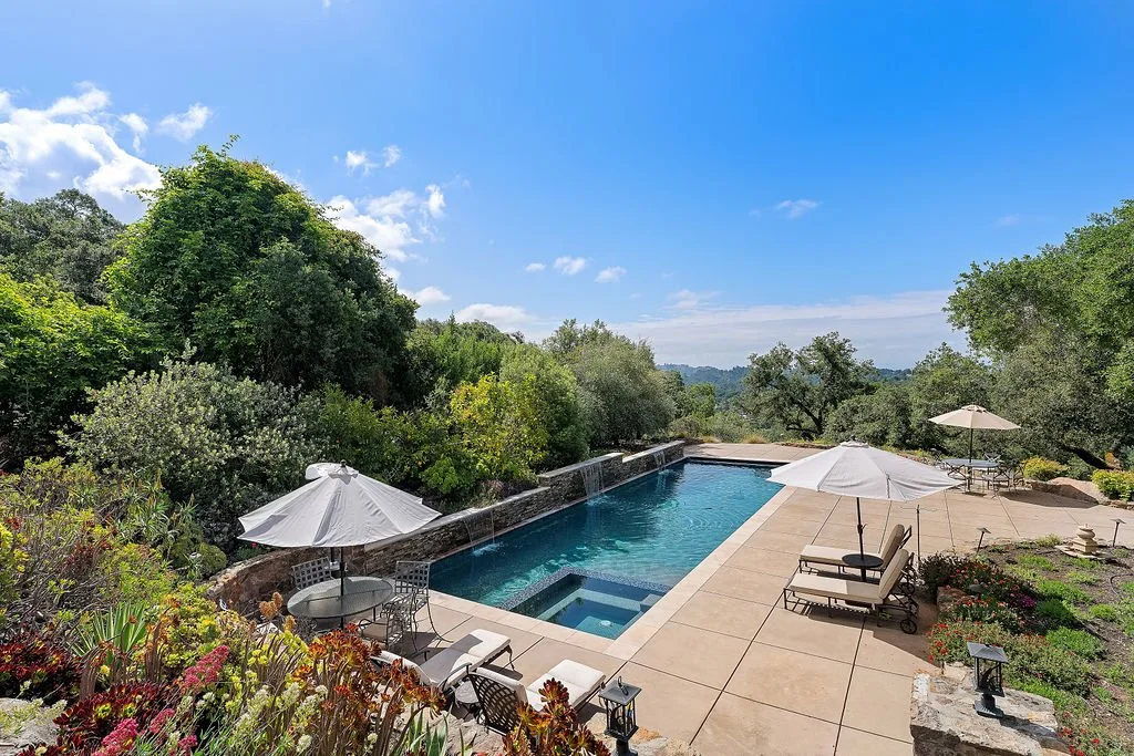 A backyard swimming pool surrounded by a stone patio with lounge chairs, umbrellas, and garden beds, overlooking a scenic landscape of trees and blue sky.