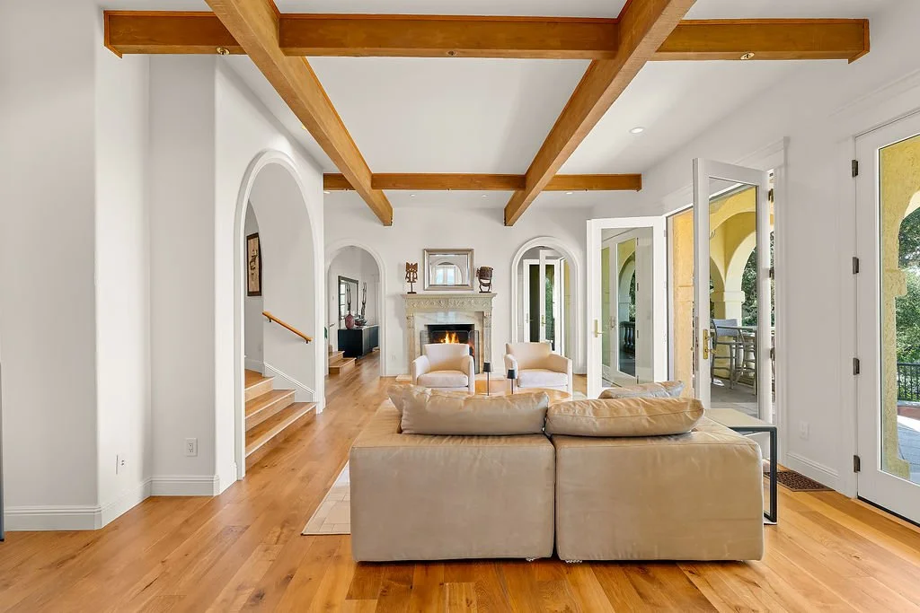 Living room with a beige sofa, two beige armchairs, a fireplace, and large glass doors leading to an outdoor patio with yellow arches.
