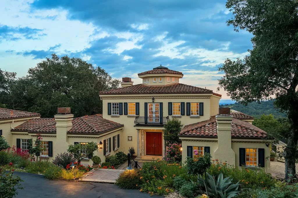 A large, elegant house with a Spanish-style terracotta roof, cream-colored stucco walls, and black shutters, set amidst lush greenery and colorful garden plants.