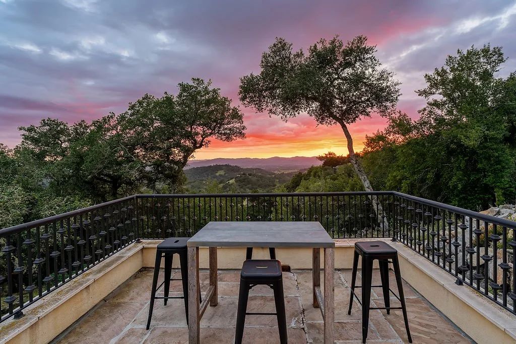A balcony with a metal railing overlooking a scenic landscape of trees and hills at sunset, with colorful pink, orange, and purple clouds in the sky.