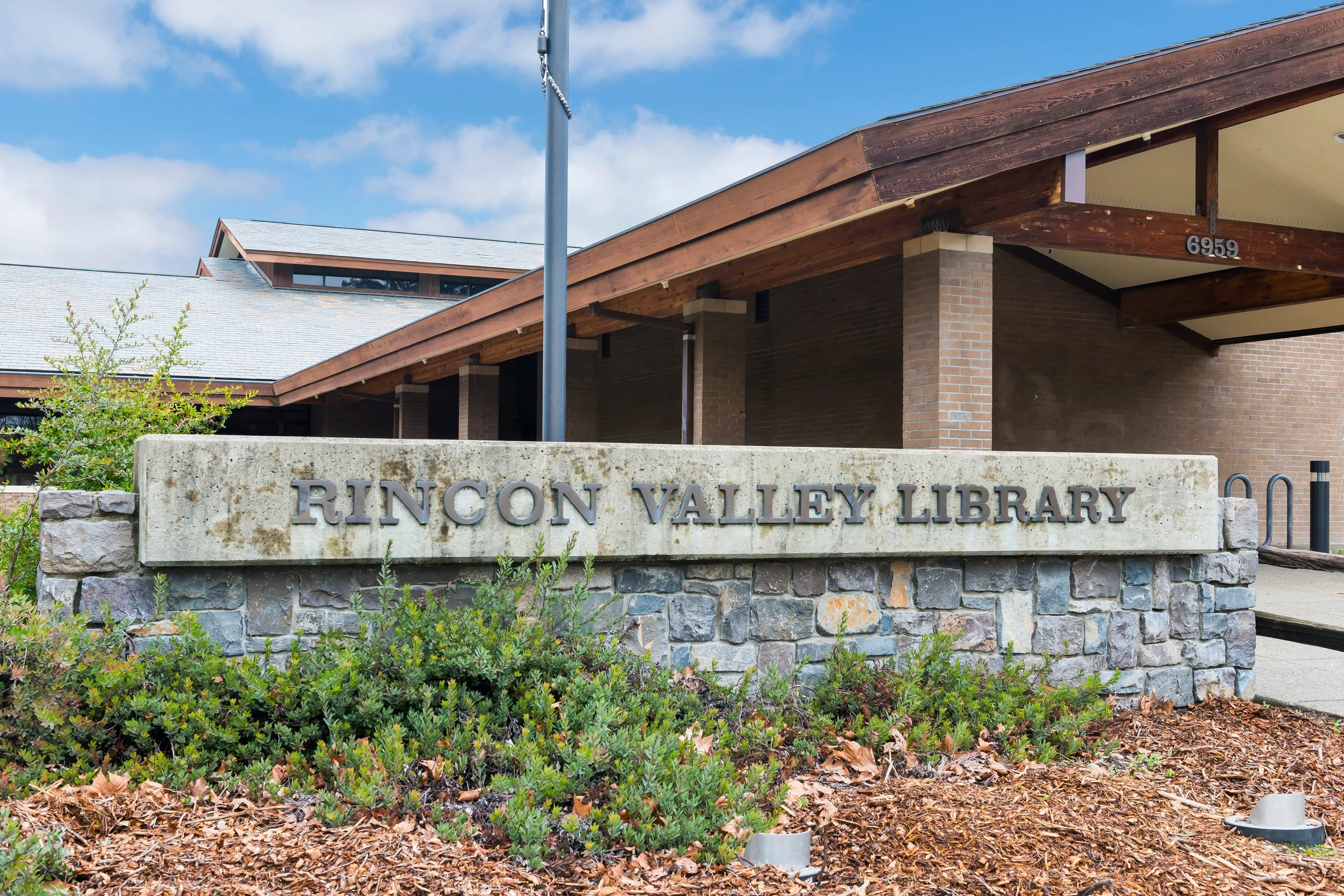 Exterior of Rincon Valley Library with a stone sign, brick building, wooden beams, and a blue sky with clouds.