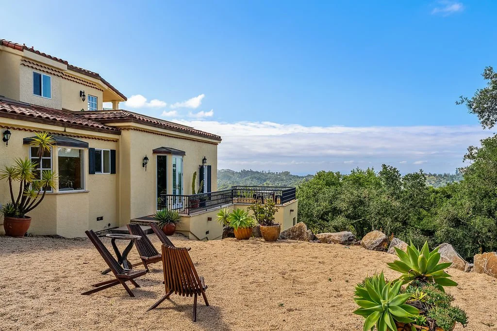A house with a beige exterior and red-tiled roof, featuring large windows, a balcony with black railing, and surrounding potted plants; outside seating with wooden chairs on sandy ground, with trees and a scenic view of distant hills and clouds in th
