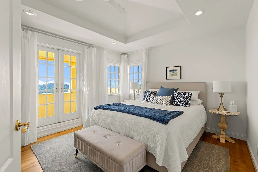 Bright bedroom with white walls, a large bed with white bedding and navy blue decorative pillows, beige upholstered headboard, small round side table with a white lamp, a window and glass door with white curtains, and a beige upholstered bench at the