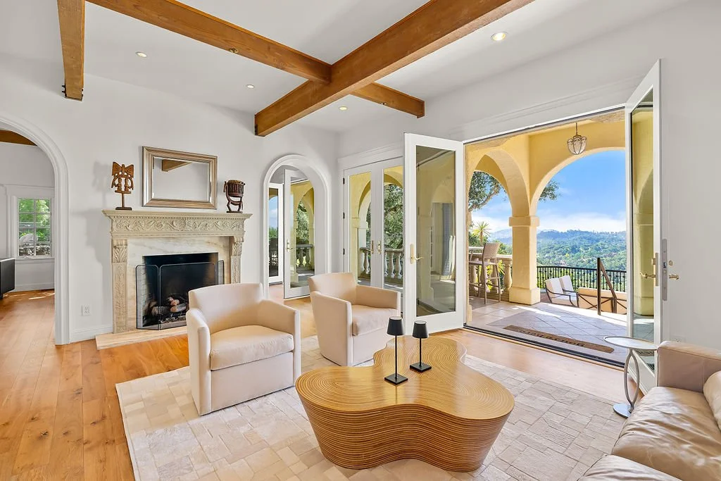 Living room with white walls, beige furniture, a stone fireplace, wooden ceiling beams, and open French doors leading to a balcony with scenic views.