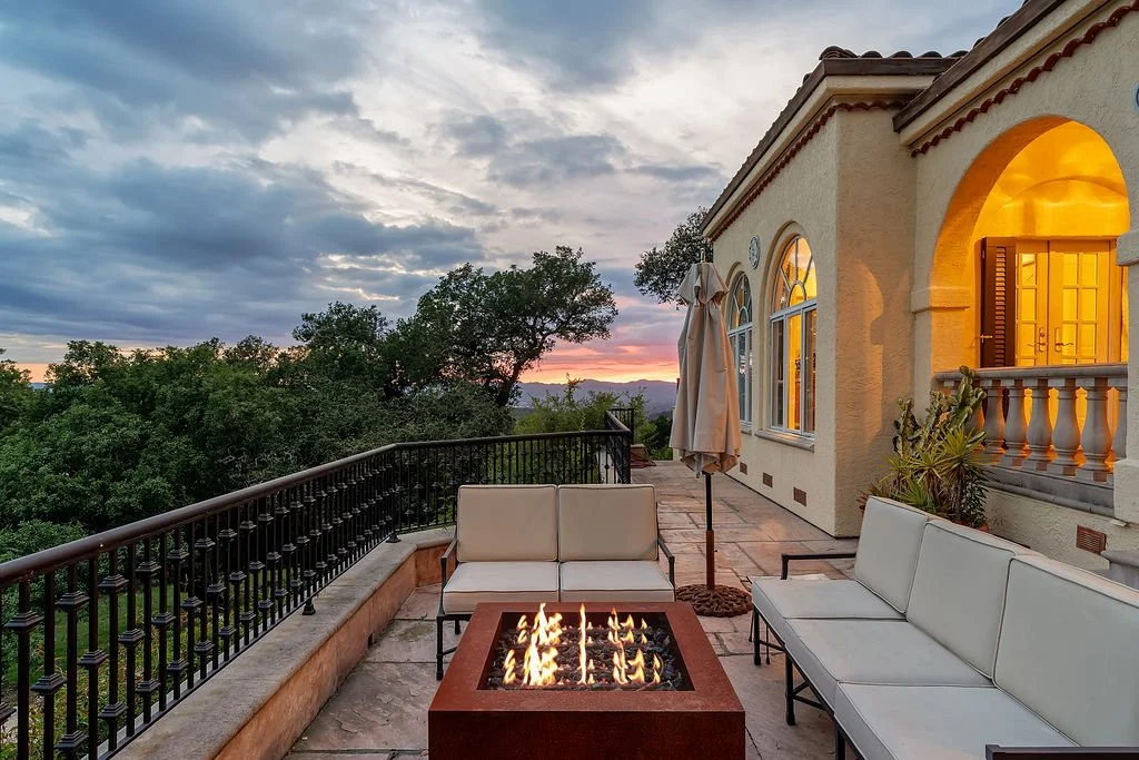 A spacious outdoor patio at sunset with a fire pit, white outdoor sofas, an umbrella, and a view of trees and a colorful sky.