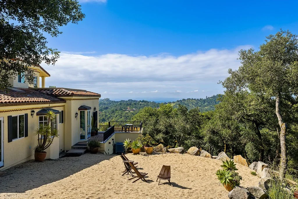 View of a hillside house with a sandy yard, potted plants, trees, and mountain scenery in the background under a blue sky with clouds.