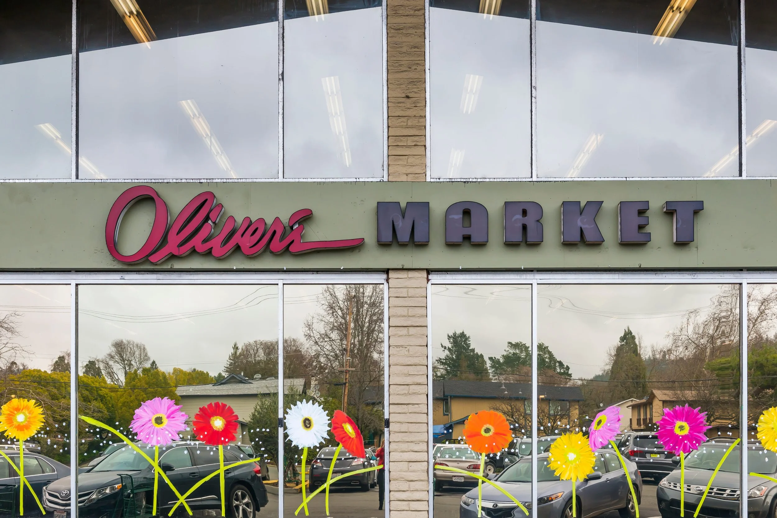 View of Oliver's Market storefront with colorful paper flowers at the window, parked cars visible outside, and reflection of trees and houses in the glass.