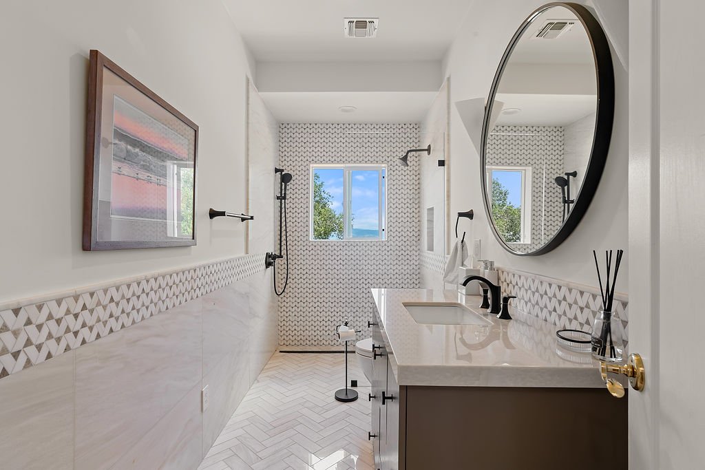 Modern bathroom with a white vanity, large round mirror, patterned backsplash, and a walk-in shower with a window.