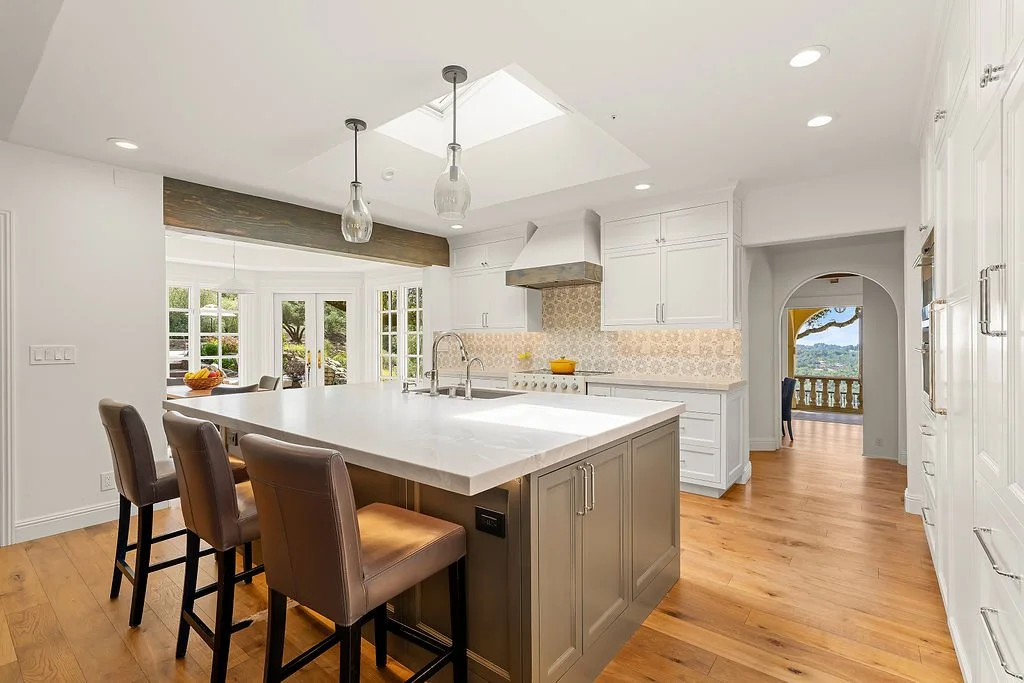 Modern kitchen with white cabinets, wooden flooring, and a large island with seating; sunlight coming through skylights and large windows.
