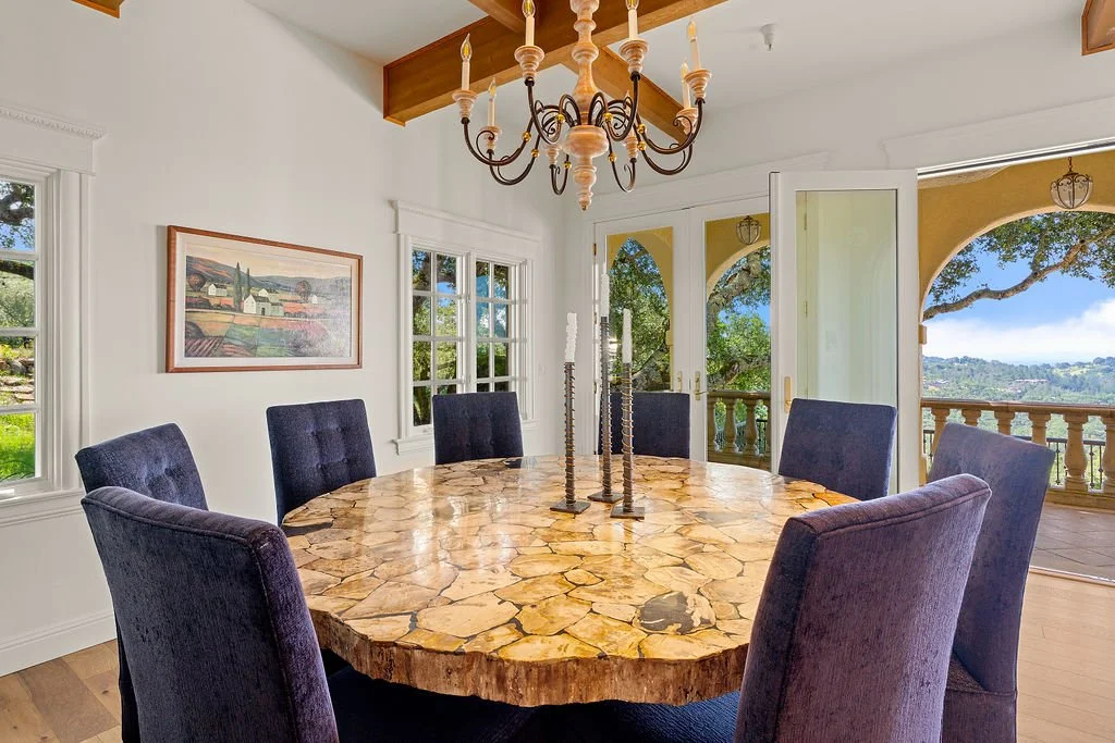 Dining room with a large round wooden table, six purple upholstered chairs, a chandelier, and a view of a balcony and green landscape outside.