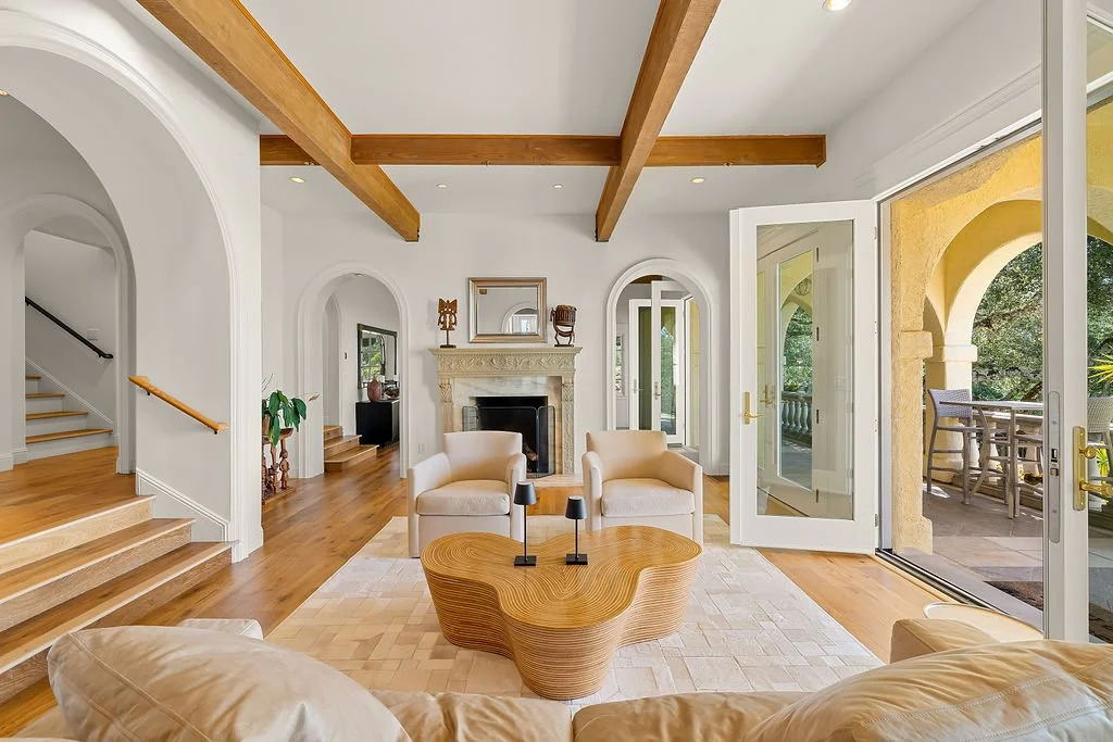 Living room with white walls, wooden beams, a fireplace, and a glass door leading to an outdoor patio with yellow arches and a furniture set.