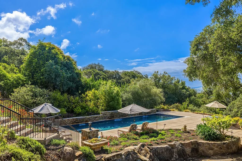 A backyard swimming pool surrounded by greenery, with lounge chairs, umbrellas, and a stone patio, under a blue sky with some clouds.