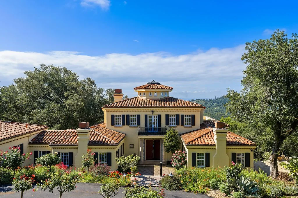 A large, Mediterranean-style house with a clay tile roof, surrounded by colorful garden plants, with trees and a blue sky in the background.