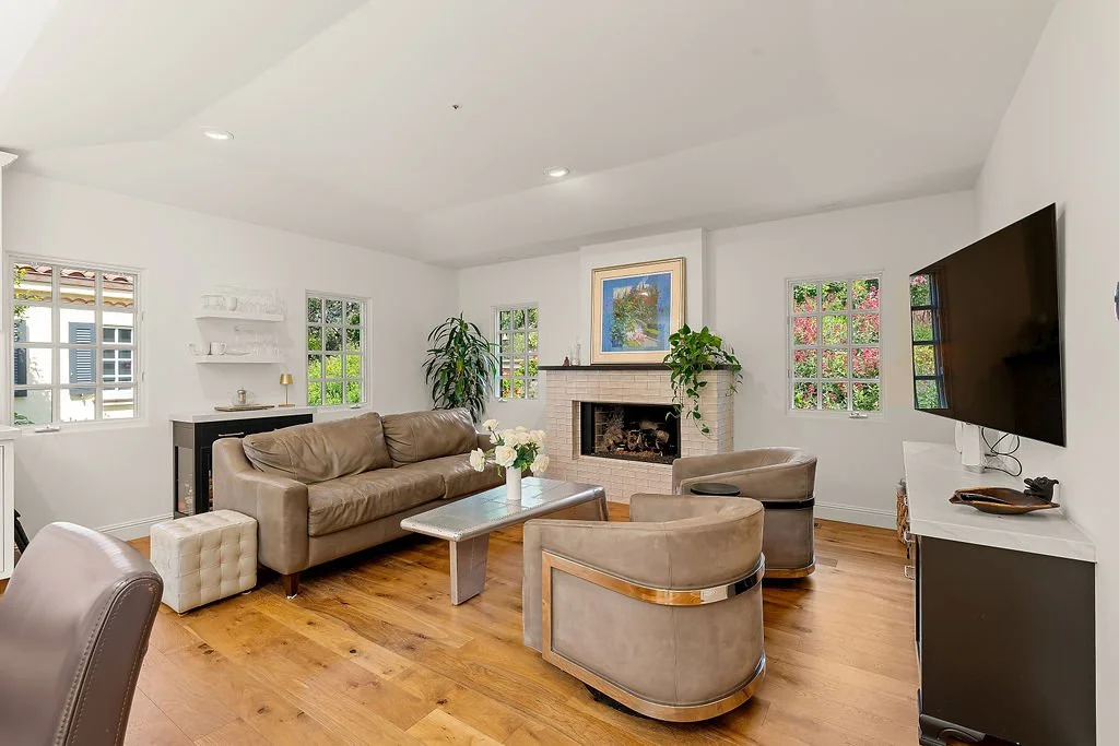 Living room with white walls, hardwood floors, a beige couch, two round beige chairs, a fireplace with a painting above, a flat-screen TV on a white and black cabinet, and windows with greenery outside.