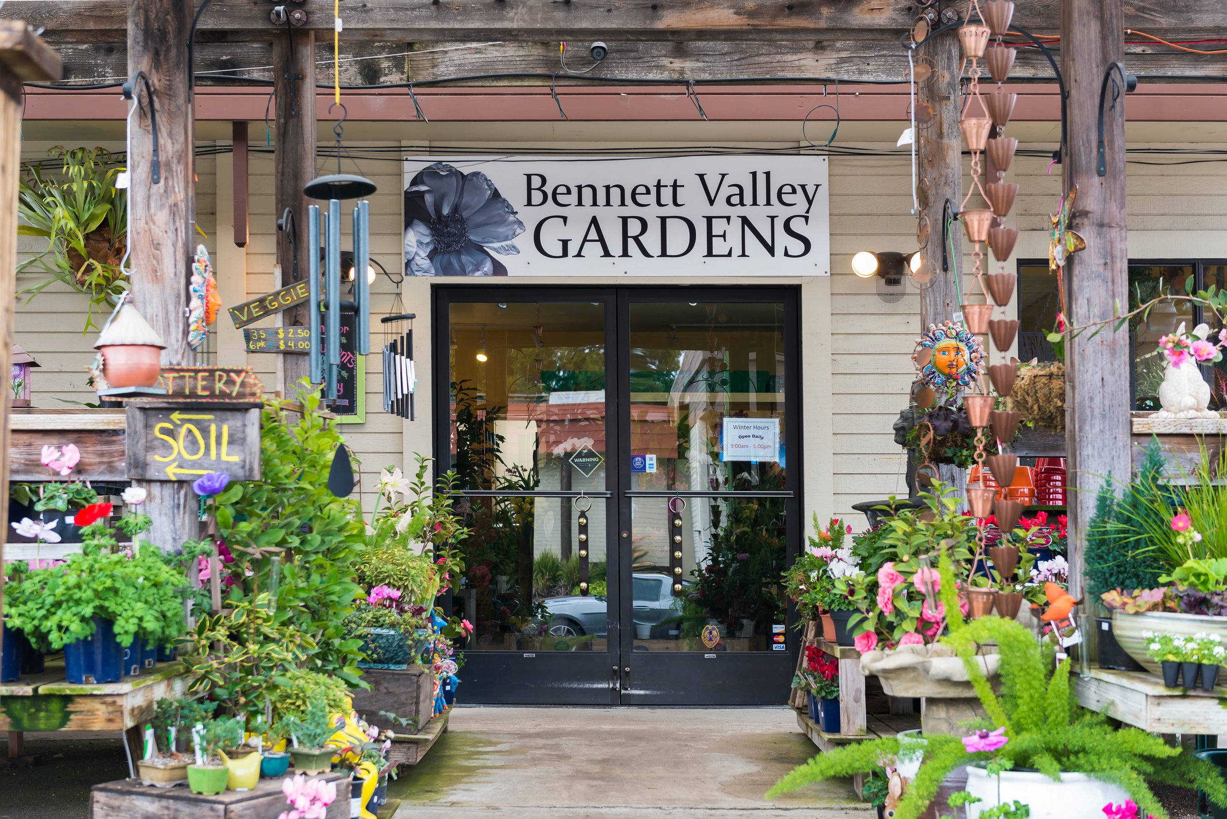 The front entrance of Bennett Valley Gardens, a plant nursery, with a sign above the door and surrounded by various potted plants, flowers, and garden decor.
