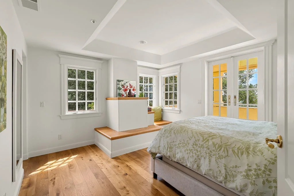 Bright bedroom with white walls, three large windows, a sliding door leading to a balcony, a bed with green leaf-patterned bedding, and wooden floors.