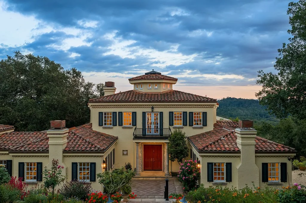 A large Mediterranean-style house with a red-tiled roof, surrounded by greenery and colorful flowers, under a partly cloudy sky during dusk or dawn.