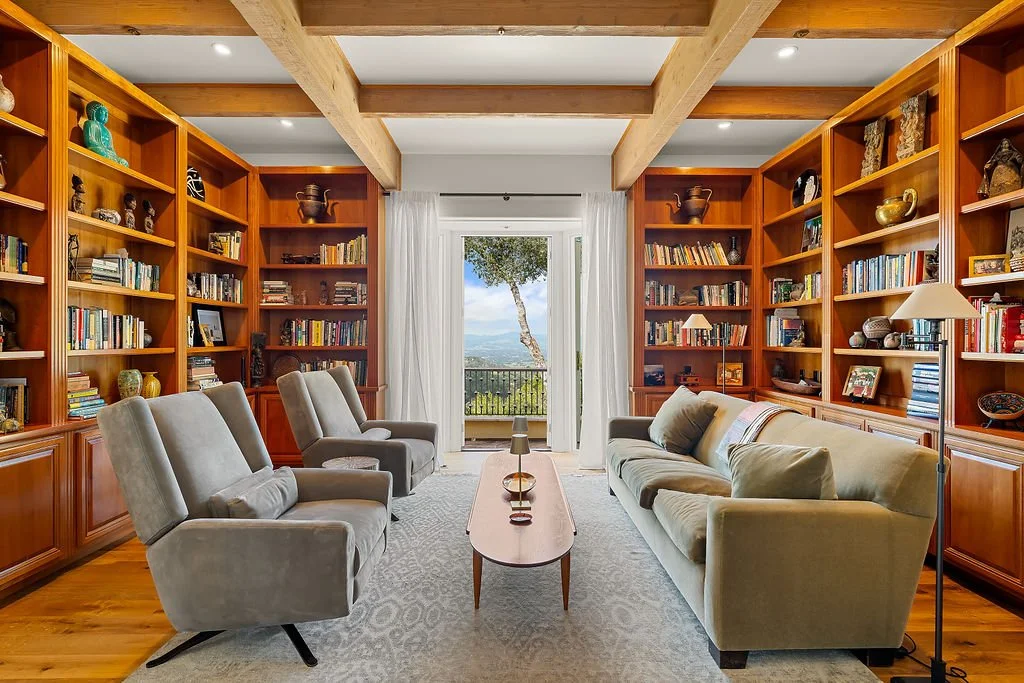 A cozy living room with built-in wooden bookshelves on all sides, a sofa and three armchairs facing each other, and a glass door opening to a balcony with a view of trees and mountains.
