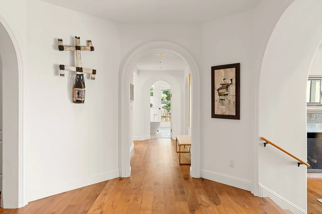 A hallway with white walls, wooden floors, and arched doorways. Decor includes indigenous wall art and a framed painting, with a bench near the wall. An open door at the end leads outside.