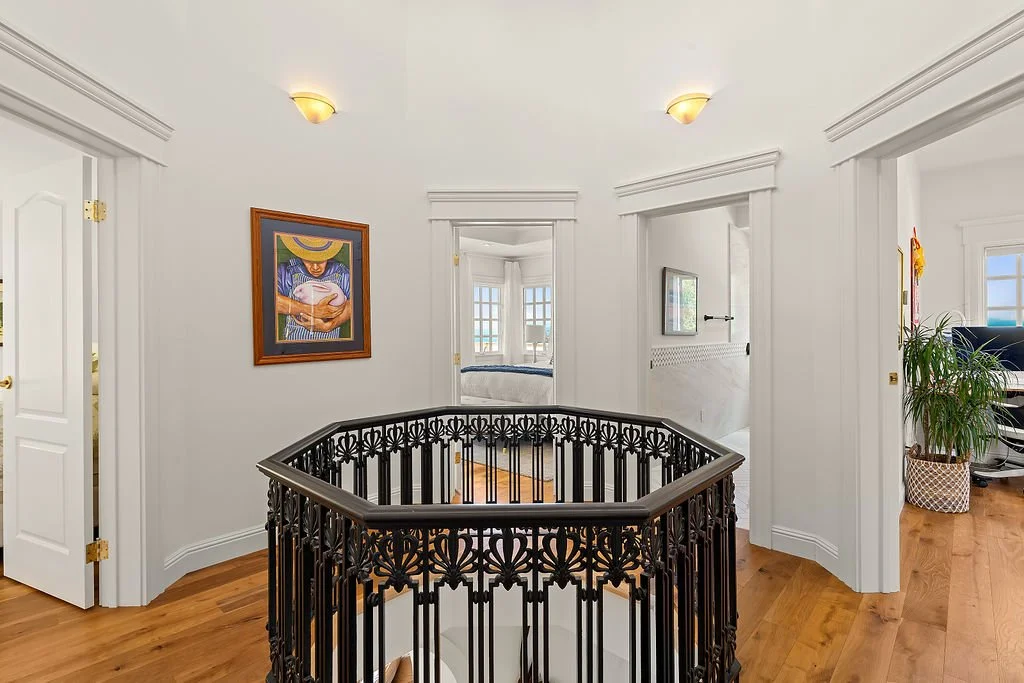 View of a bright hallway with white walls and wooden floors, featuring a decorative black metal railing around a staircase opening, framed artwork on the wall, and doorways leading to other rooms with bay windows and a potted plant.