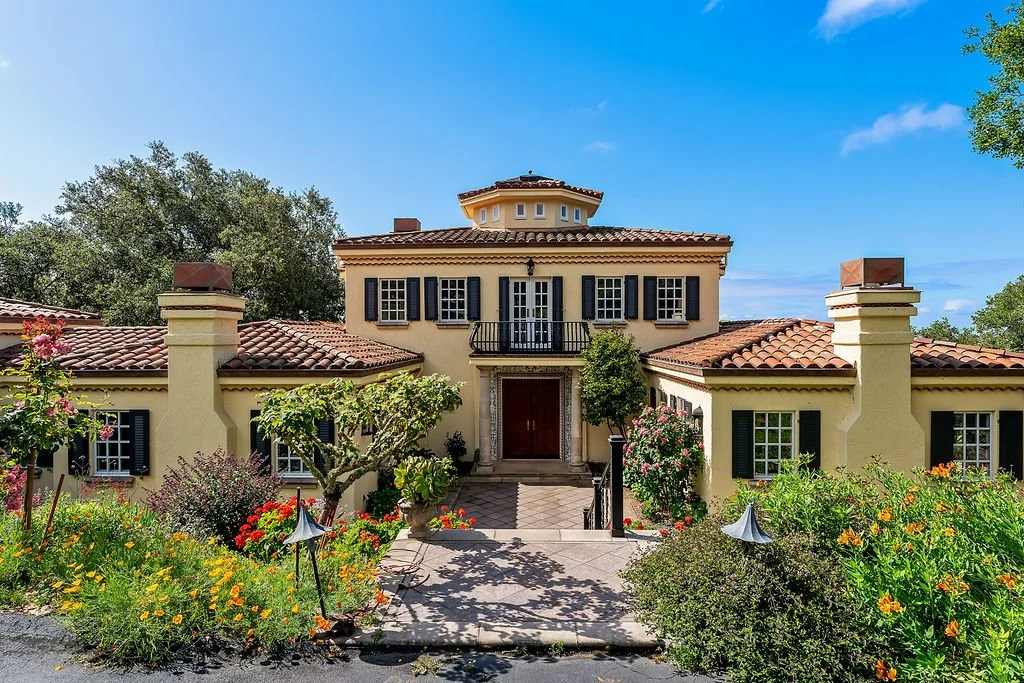 A large Mediterranean-style house with a terracotta tiled roof, beige stucco walls, black shutters, and a small balcony above the front entrance, surrounded by a colorful garden with various bushes and flowers under a clear blue sky.