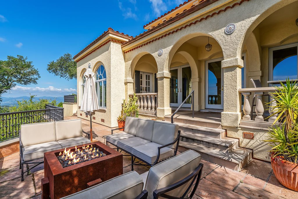 Outdoor patio area with beige cushioned seating and a fire pit, attached to a cream-colored stucco house with arched doorways, large windows, potted plants, and a scenic view of trees and sky.