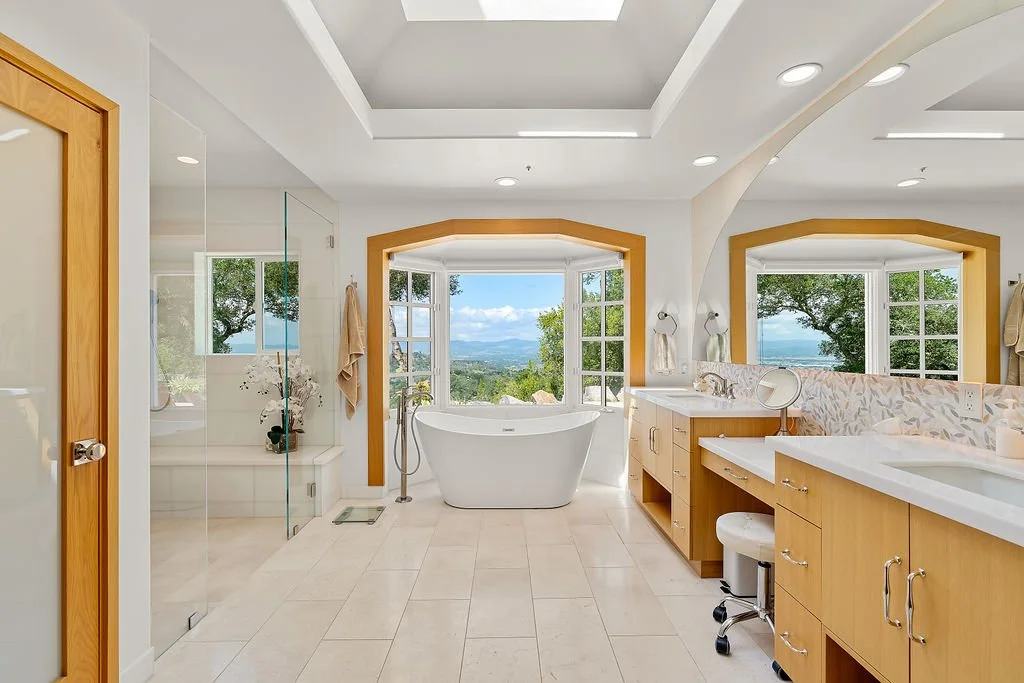 Bright bathroom with a white freestanding bathtub in front of large windows showcasing a scenic view of trees and mountains.