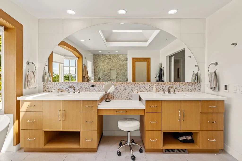 Double vanity bathroom with large mirror, wooden cabinets, white countertops, and a stool in the center. Towel racks with towels and a window with greenery outside.