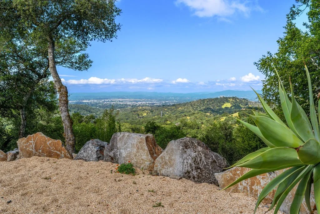 A scenic view from a hillside with sandy ground, large rocks, green trees on either side, and a distant view of rolling hills, a valley, and a city under a partly cloudy sky.