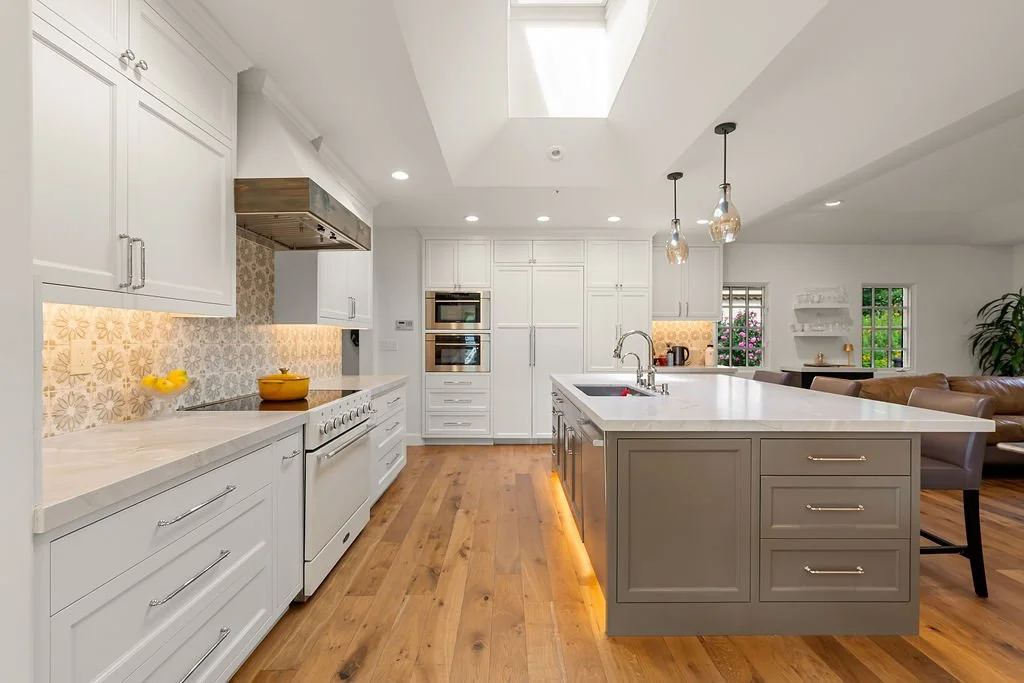Modern kitchen with white cabinets, a gray island, pendant lighting, and hardwood floors, with a living room area in the background.