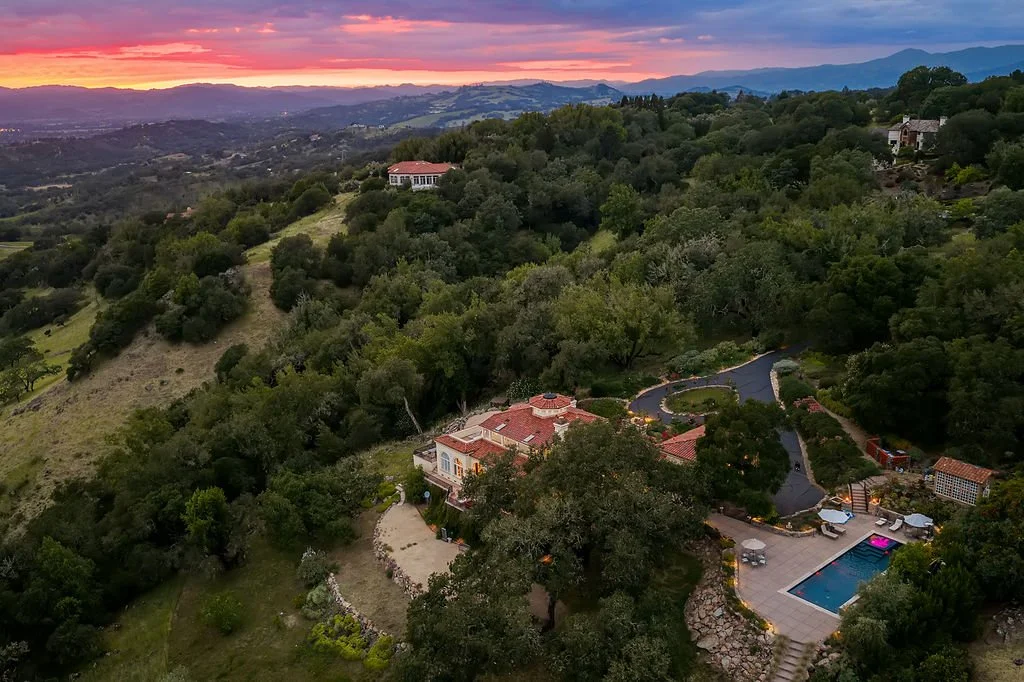 Aerial view of a hillside property with a swimming pool, a winding driveway, and several houses surrounded by lush green trees during sunset with mountains in the background.