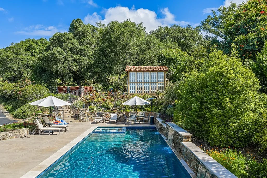 View of a backyard swimming pool with lounging chairs, umbrellas, and a garden with trees and shrubs, and a small structure with large windows in the background.