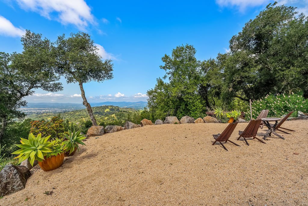 Outdoor patio with three wooden lounge chairs, a small table, potted succulent plants, and a scenic view of trees, mountains, and a blue sky with clouds.