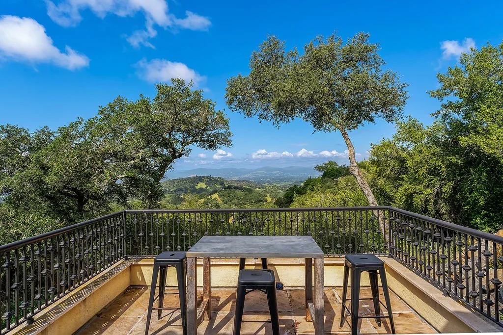 Outdoor patio with black metal railings, three black stools, and a gray table overlooking a lush green landscape with trees and distant hills.