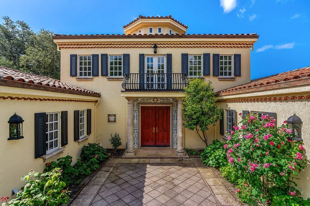 Front view of a Mediterranean-style house with a red door, yellow stucco walls, black shutters, and a small balcony, surrounded by greenery and blooming pink flowers.