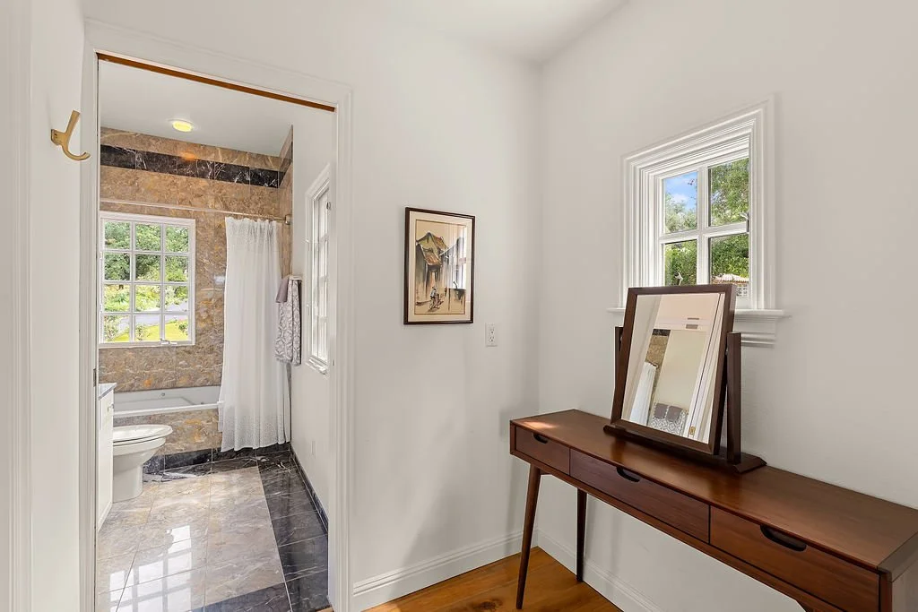 View of a small hallway leading into a bathroom with marble tiles, a window with a view of green trees, and a white toilet. The hallway has a wooden console table with two framed mirrors and a window showing a bright, sunny day outside.