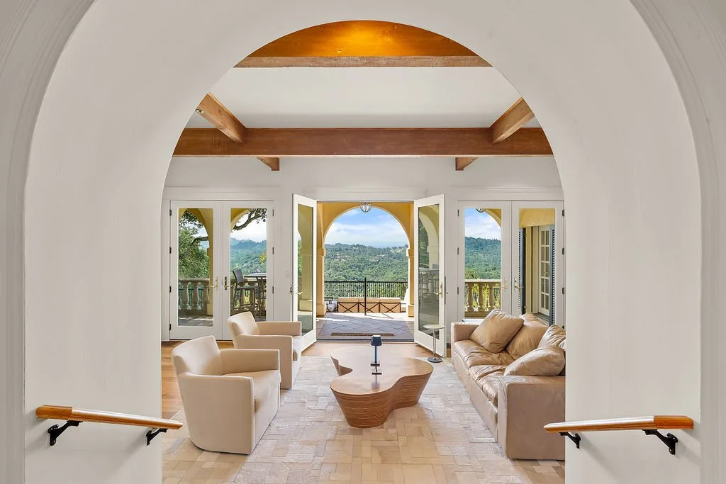 Living room with beige sofas and chairs, wooden coffee table, open doors leading to a balcony with scenic mountain view.