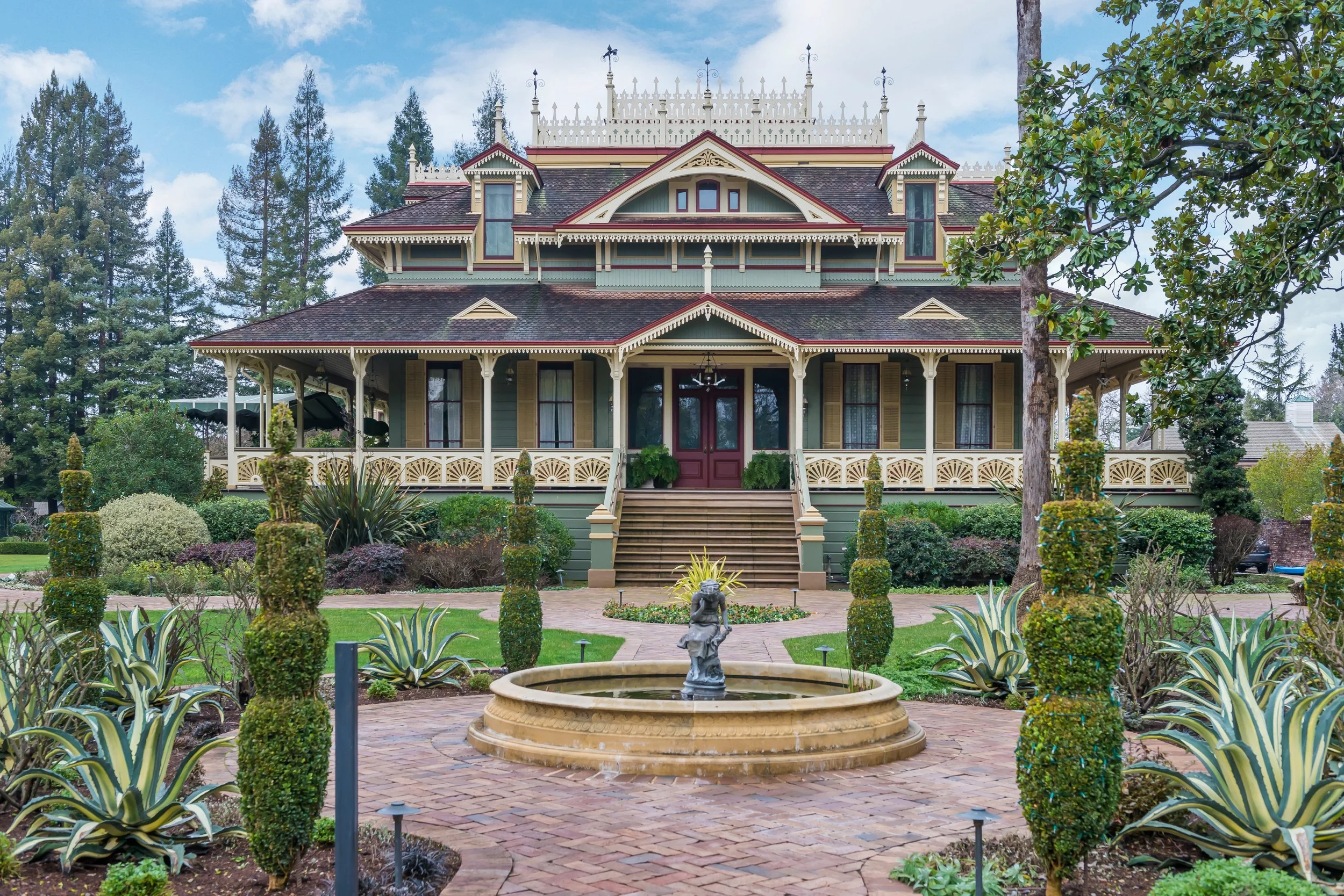 A Victorian-style house with a front porch, surrounded by a landscaped garden with bushes, trees, and a fountain in the foreground, under a partly cloudy sky.