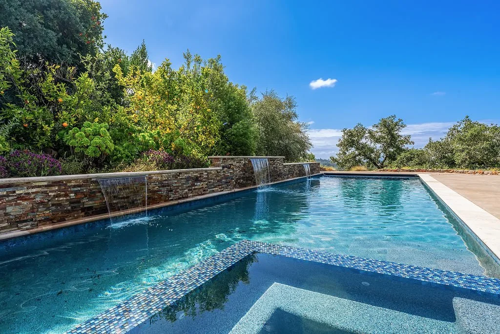 A luxurious outdoor swimming pool with a stone waterfall feature, surrounded by lush green trees and a clear blue sky.