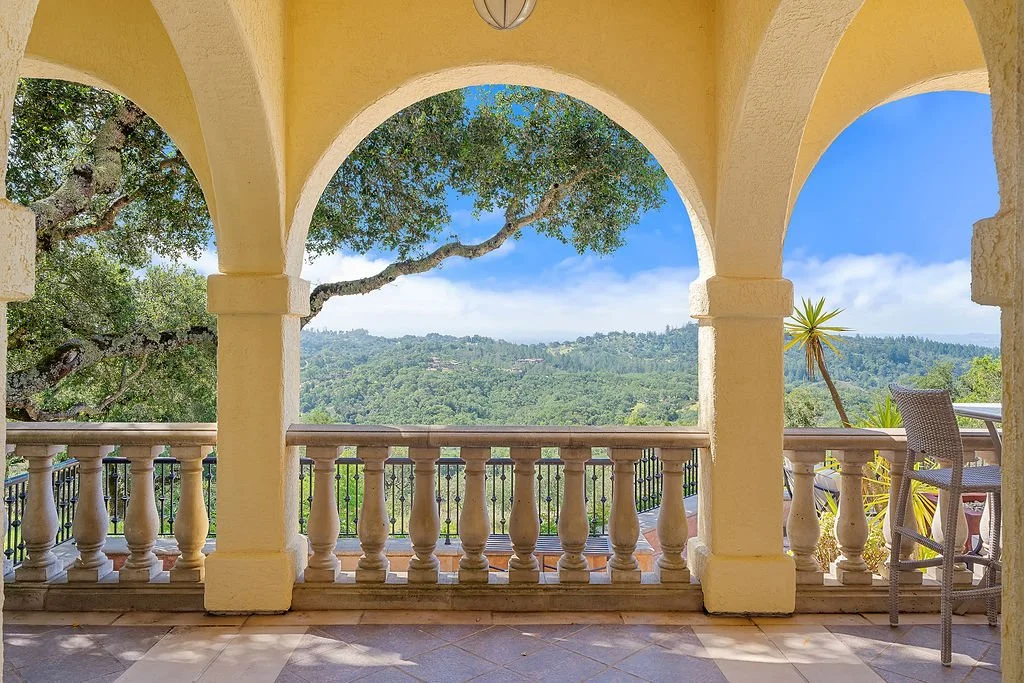 View from a balcony with yellow arches and railing, overlooking green hills and trees with a cloudy blue sky.