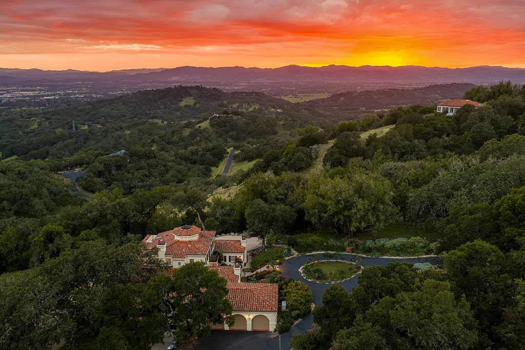 A scenic sunset over a hilly, green landscape with scattered trees and homes, featuring a large house with a red tile roof and a winding driveway.