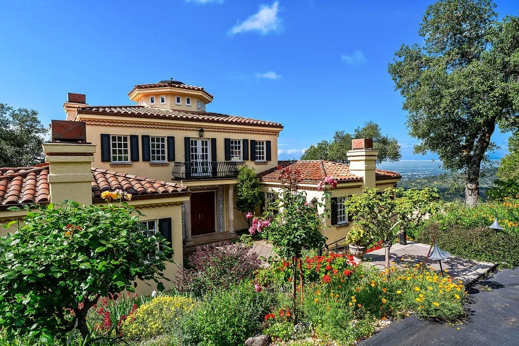 A Mediterranean-style house with a tiled roof, surrounded by lush plants and colorful flowers, set against a backdrop of a bright blue sky and green trees.