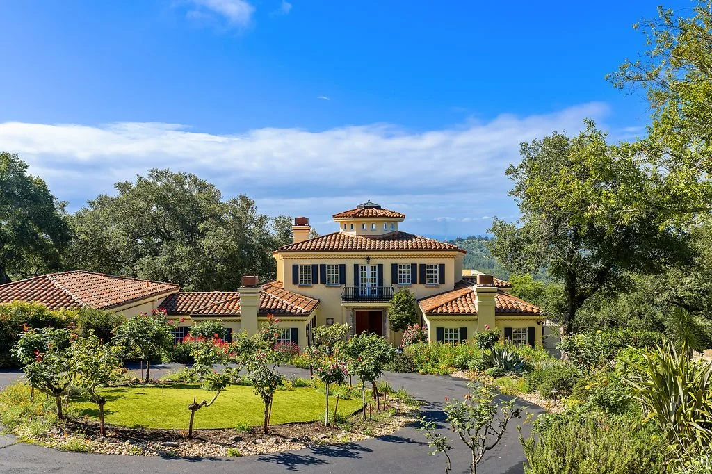 A large house with a tiled roof surrounded by a garden with trees and bushes, under a partly cloudy sky.