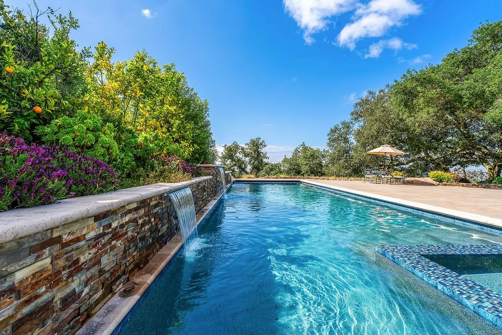 A swimming pool with a brick waterfall feature on the left, surrounded by lush green trees and plants, with a patio area including an umbrella and chairs in the background, under a clear blue sky.